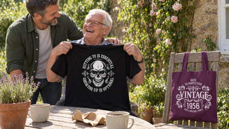 Grandad laughing on a garden patio holding up a Hitting 70 is Essential T-shirt, a plum Vintage 1956 tote on a chair