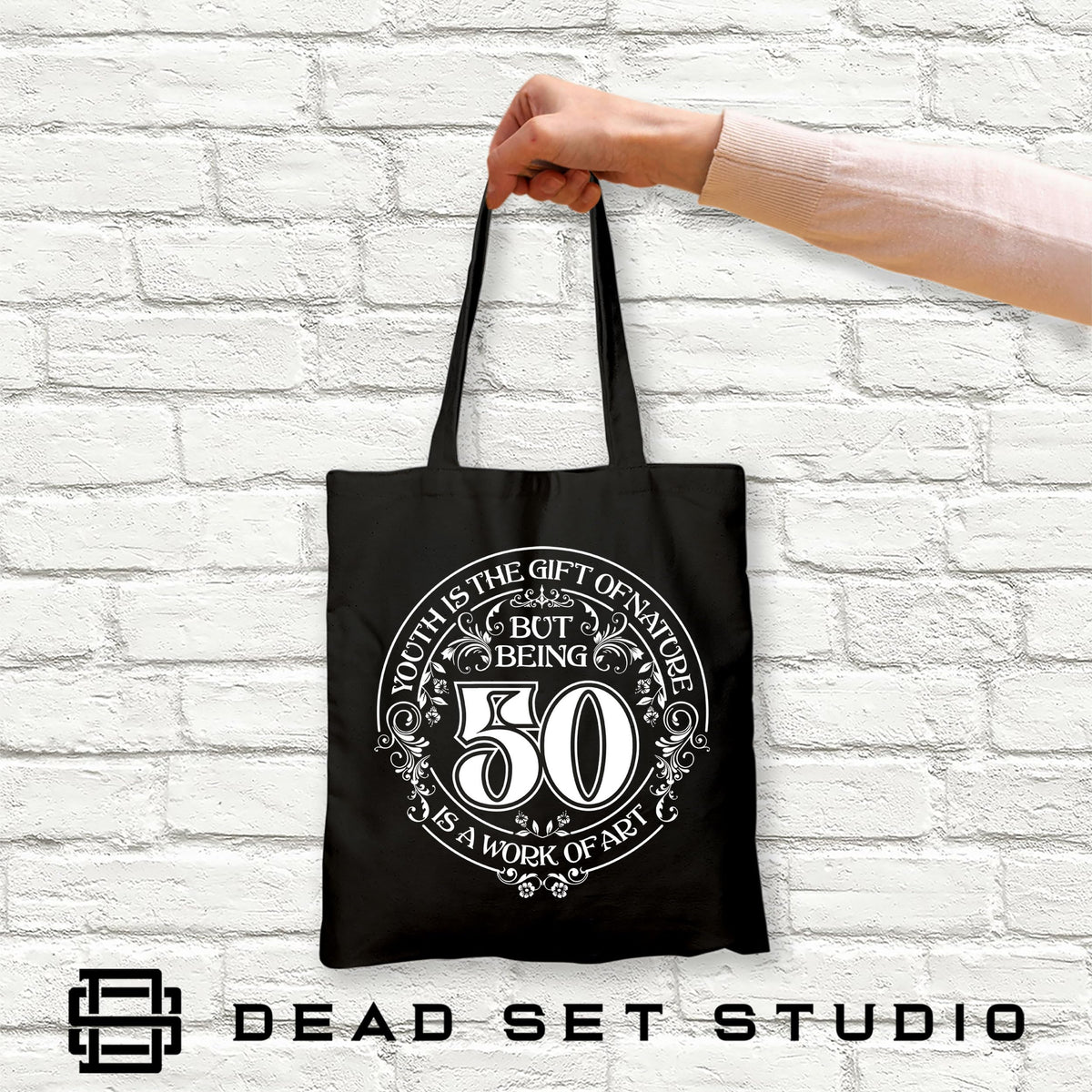 Hand holding the black Being 50 is a Work of Art 50th birthday tote bag against a white brick wall