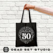 Hand holding the black Being 50 is a Work of Art 50th birthday tote bag against a white brick wall
