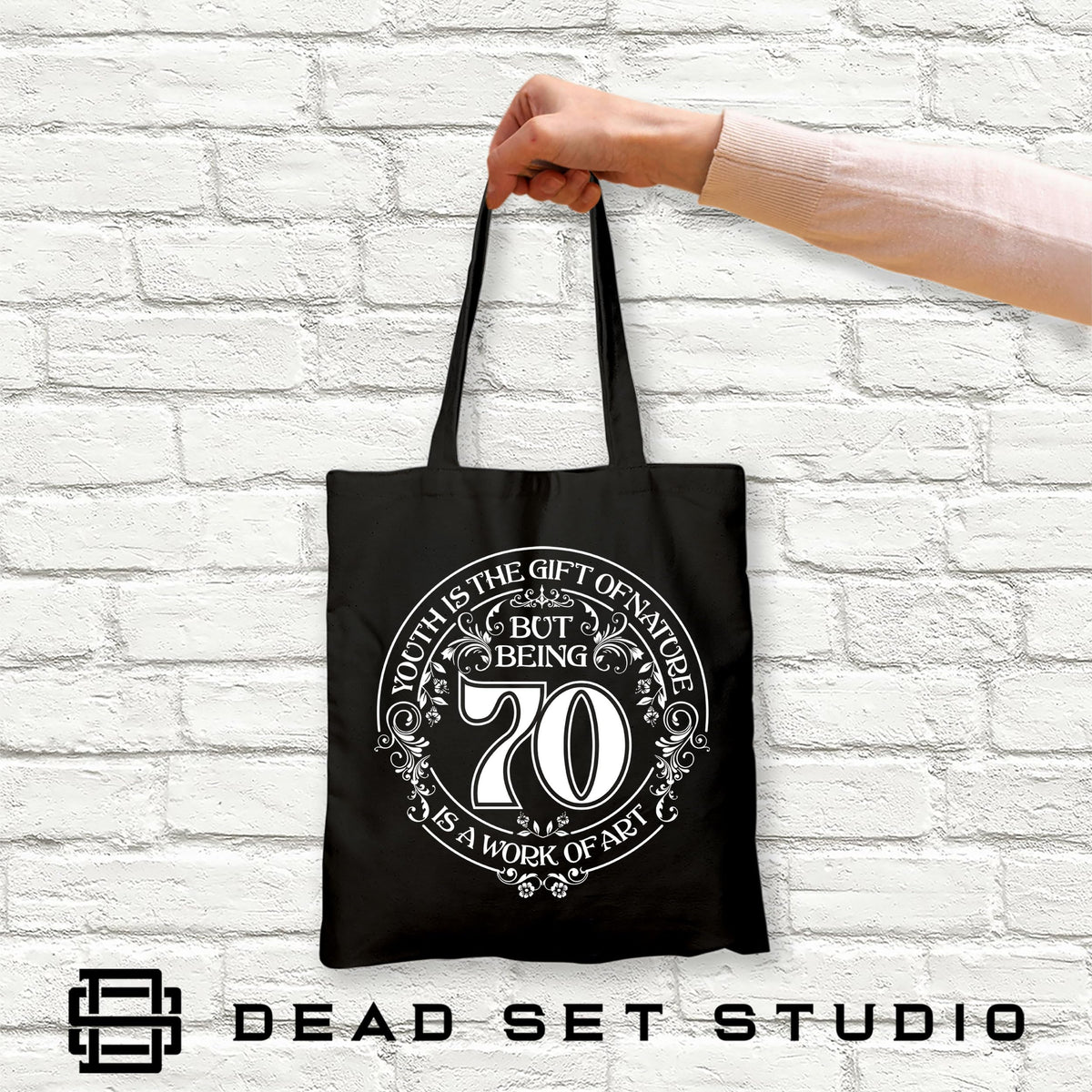 Hand holding the black Being 70 is a Work of Art 70th birthday tote bag against a white brick wall