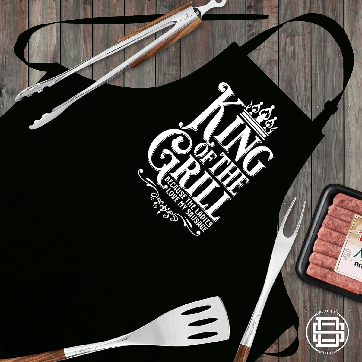 King of the Grill black apron laid flat with tongs, spatula and sausages