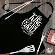 King of the Grill black apron laid flat with tongs, spatula and sausages