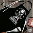 Manly Man skull apron laid flat with BBQ tongs, fork, spatula and sausages