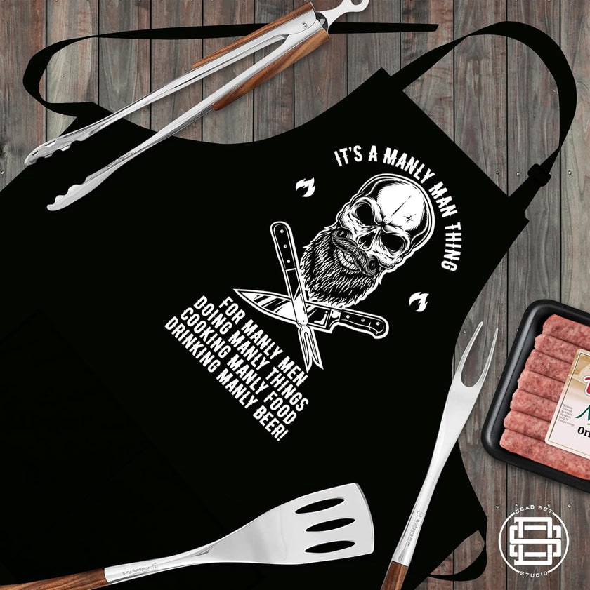 Manly Man skull apron laid flat with BBQ tongs, fork, spatula and sausages