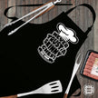 Never Trust a Skinny Chef black apron laid flat with tongs, fork, spatula and sausages