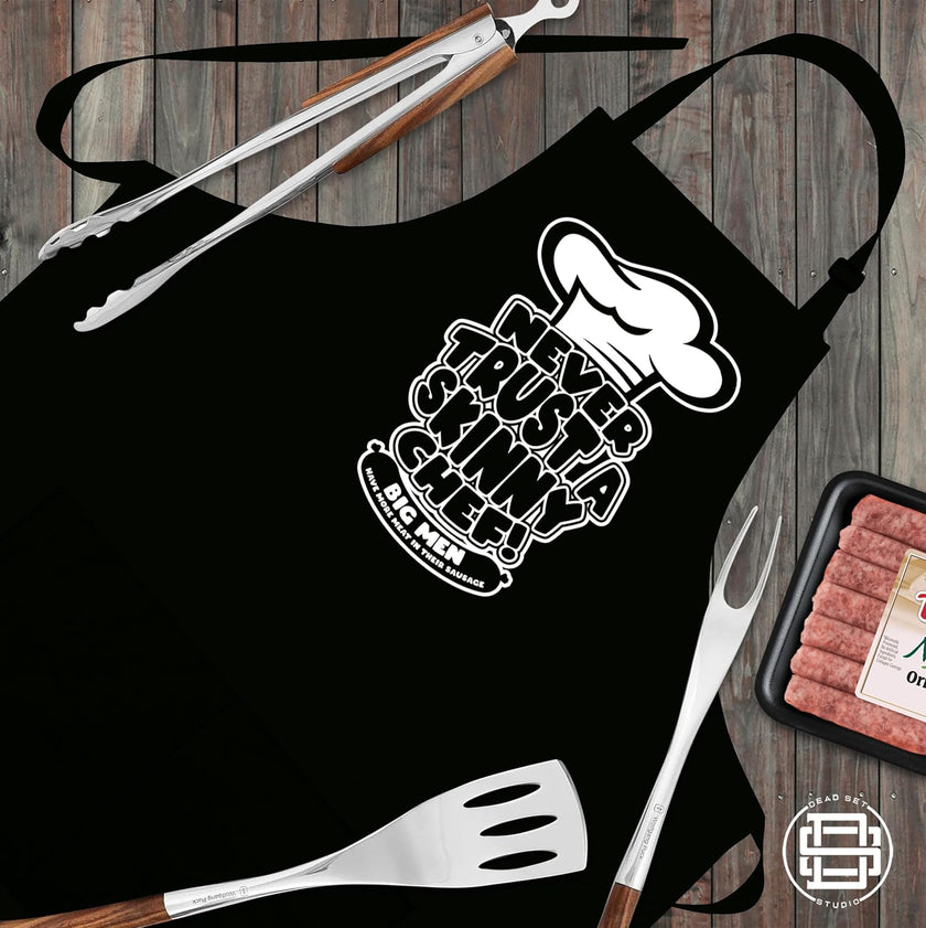 Never Trust a Skinny Chef black apron laid flat with tongs, fork, spatula and sausages