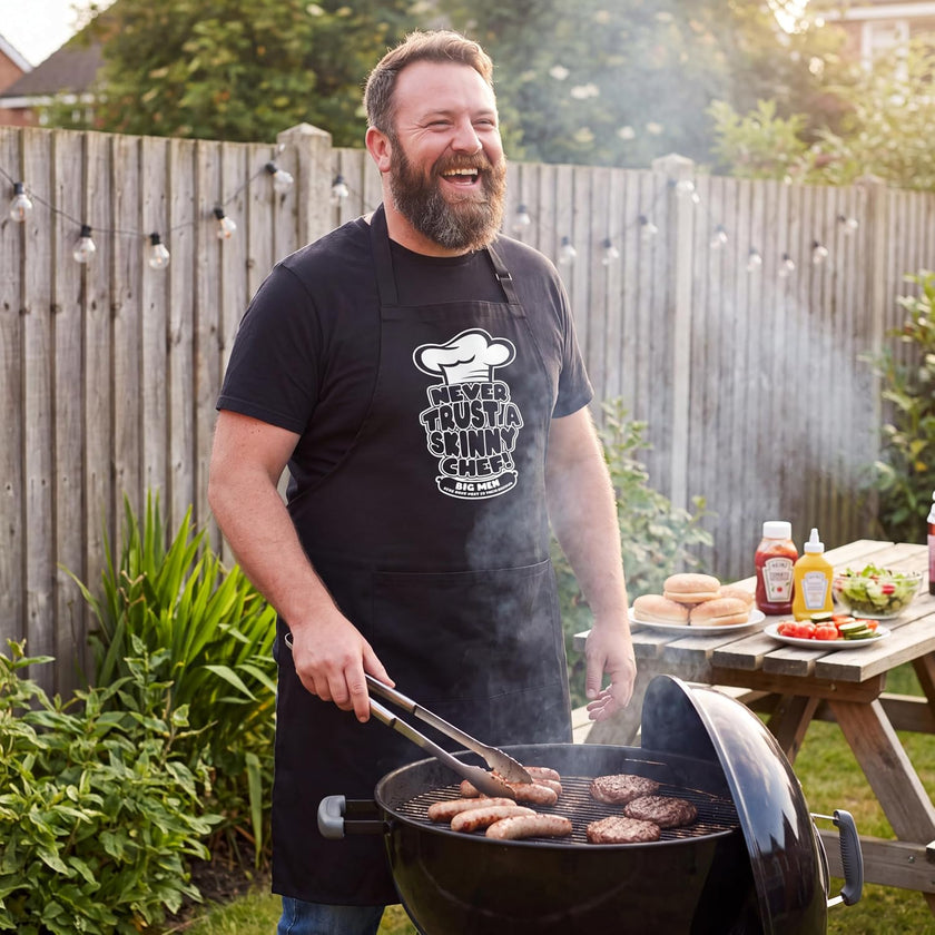 Bearded man wearing the Never Trust a Skinny Chef apron grilling sausages in the garden